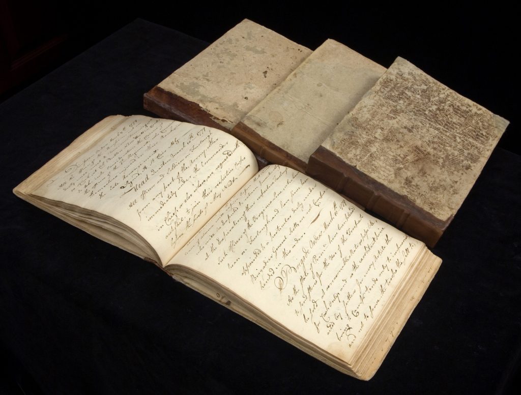 Four manuscript orderly books from the Revolutionary War, one open with handwriting filling the pages and three others closed beside it