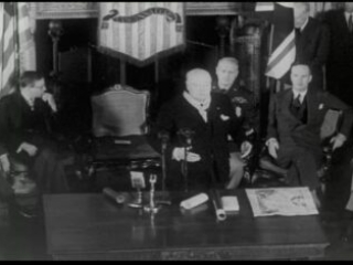 Click for a larger view. Black-and-white image of Winston Churchill giving a speech in front of a fireplace with men seated behind him