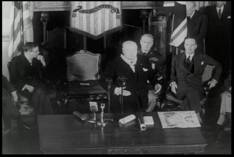 Black-and-white image of Winston Churchill giving a speech in front of a fireplace with men seated behind him