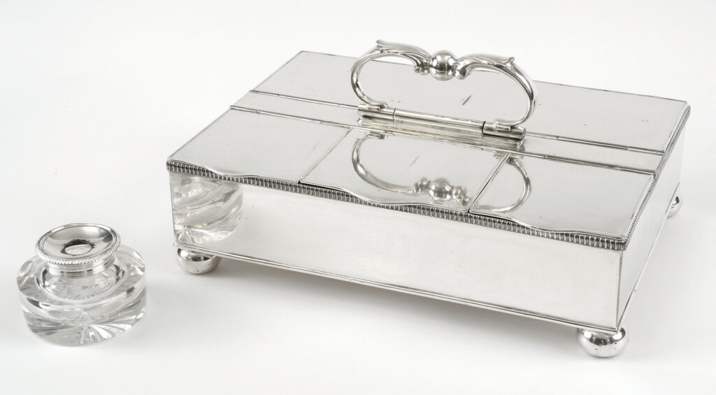 Rectangular silver box with a handle next to a glass and silver inkwell